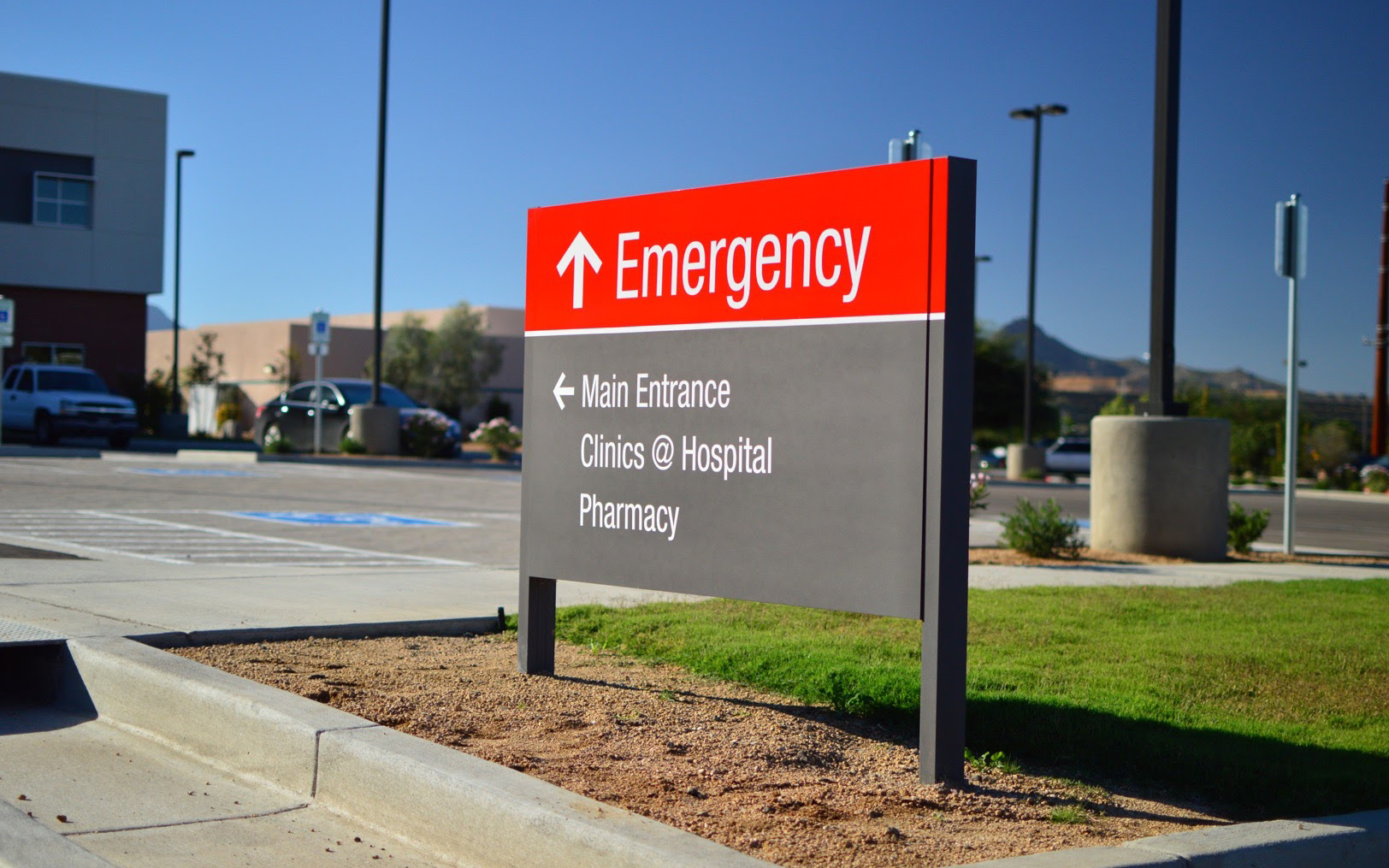 Exterior post-and-panel sign directing visitors to emergency services, the main entrance, clinics and pharmacy
