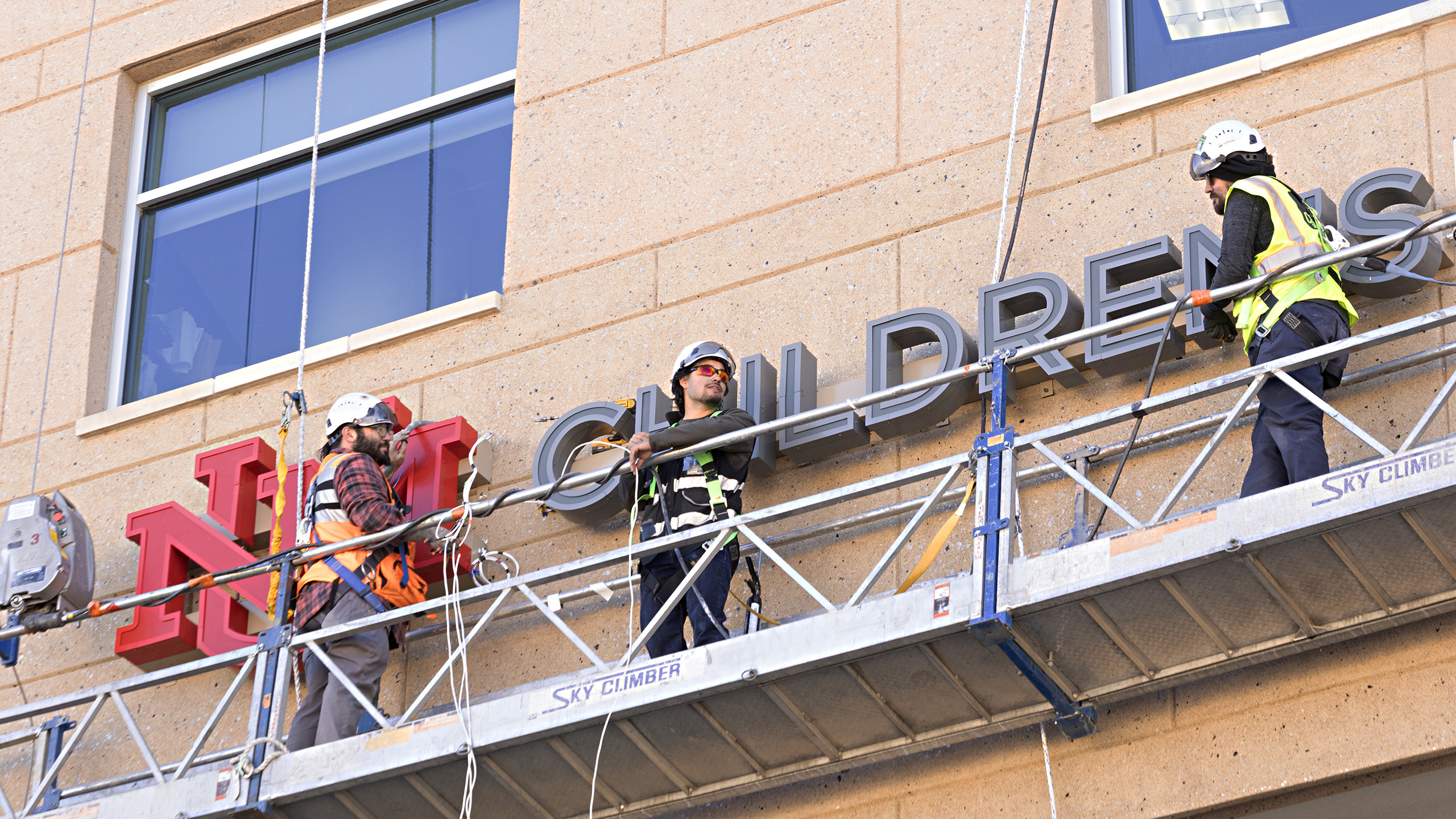Close-up of sign installers placing face illuminated channel letters on the UNM Children's Hospital Pavilion exterior.