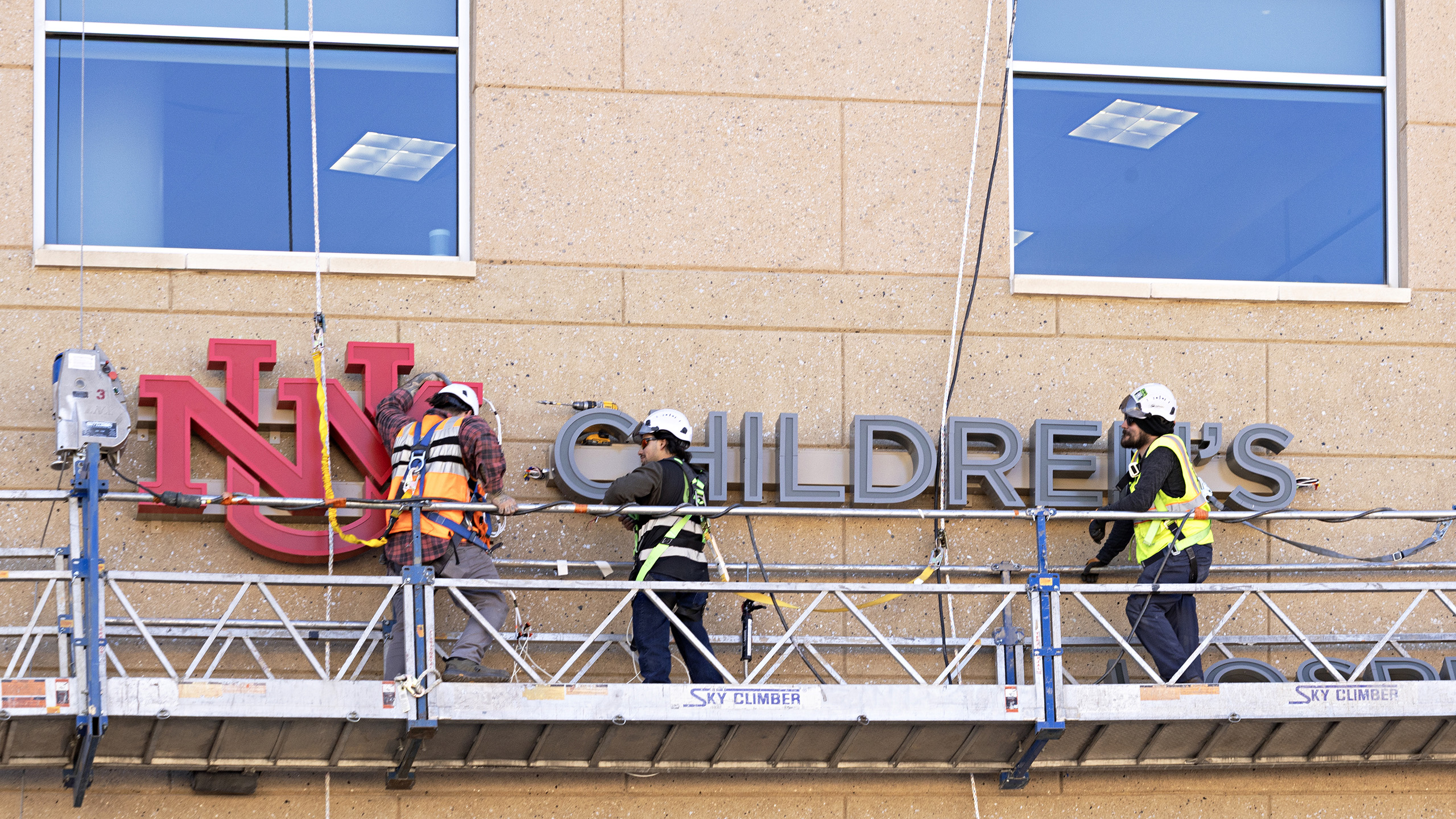 Sign installers working from suspended scaffolding to install illuminated channel letters at UNM Children's Hospital Pavilion.