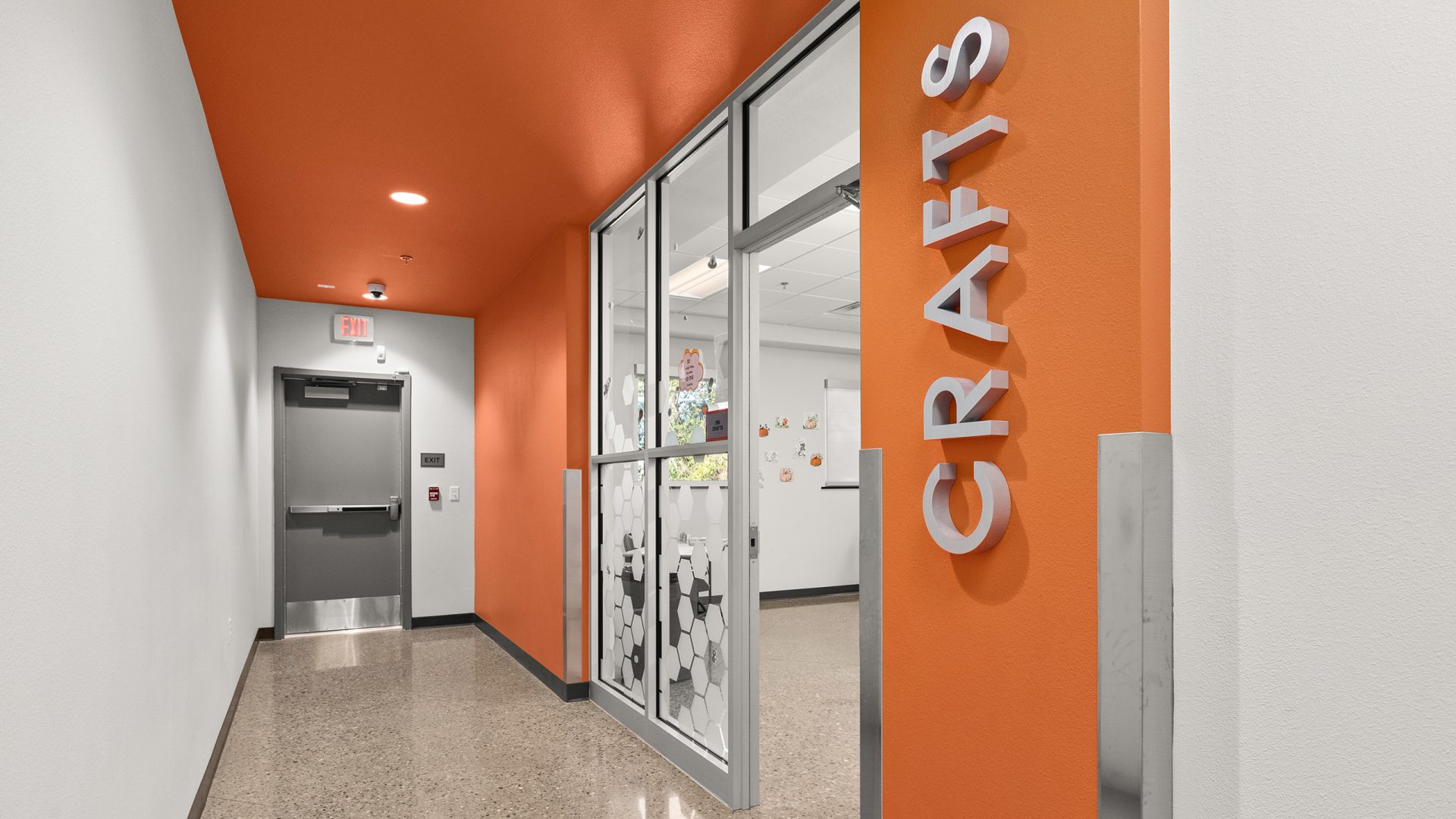 Brushed satin cast aluminum dimensional letters spelling Crafts mounted vertically on an orange wall near glass entry doors.