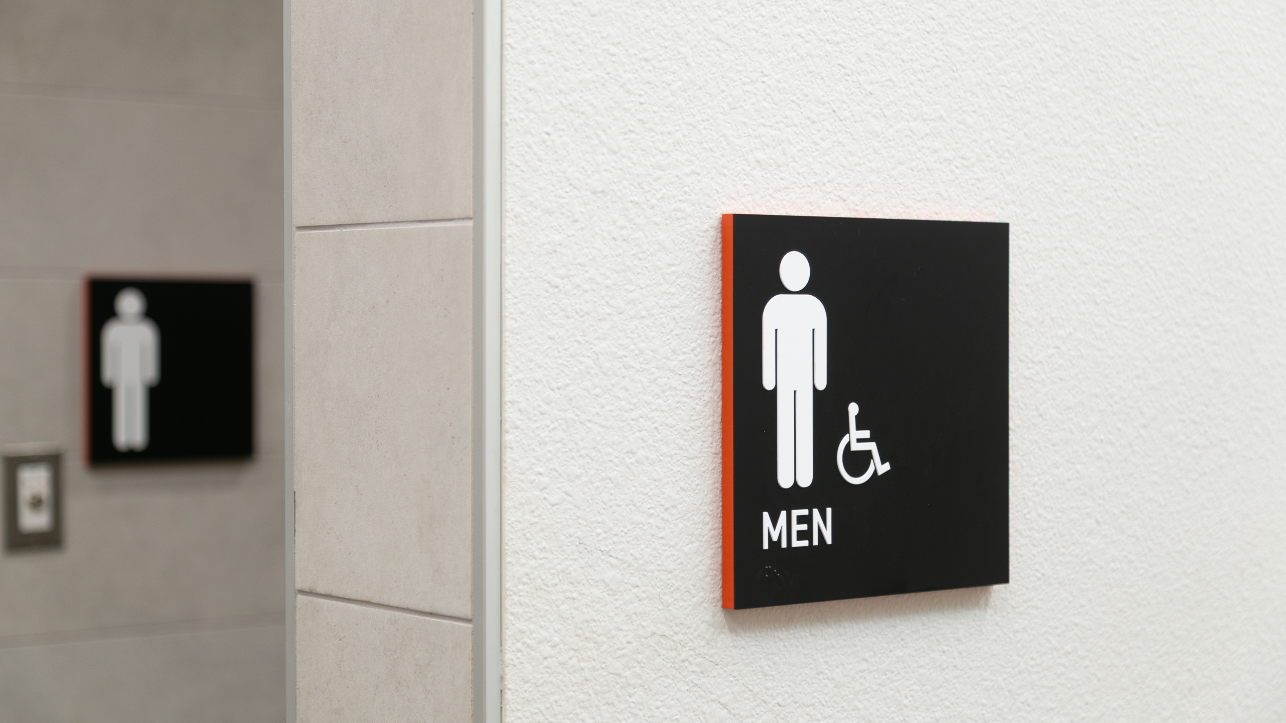 Black ADA men’s restroom sign with white pictogram, tactile text and Braille