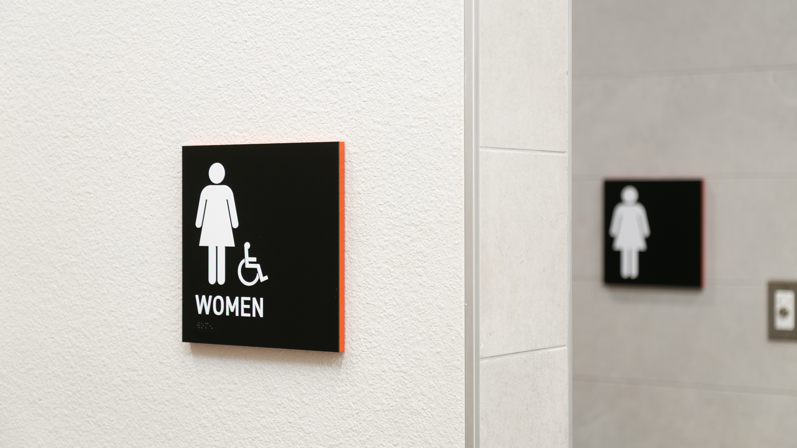 Black ADA women’s restroom sign with white pictogram, tactile text and Braille